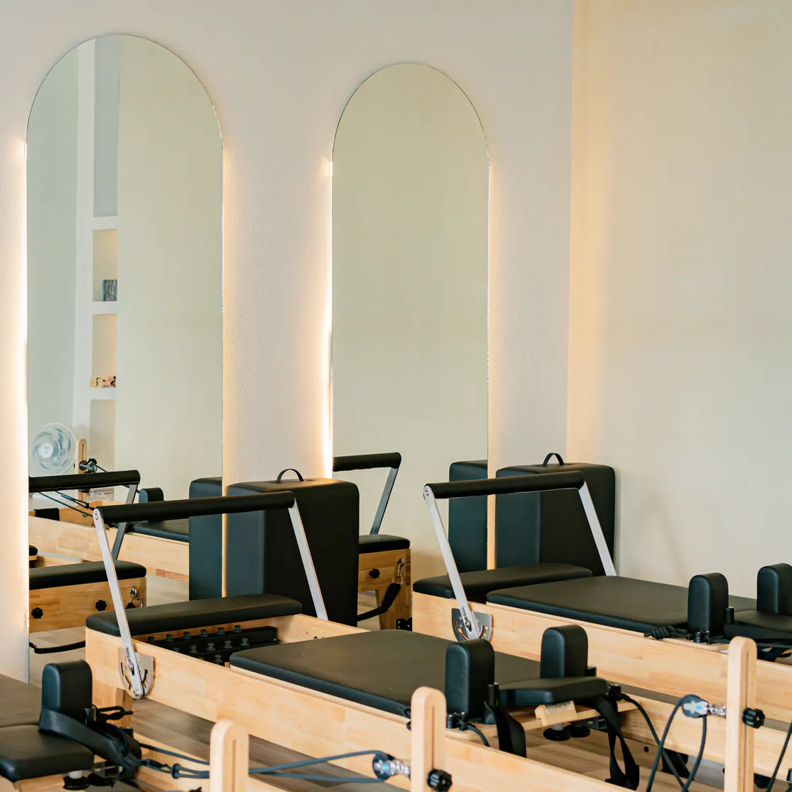 Sala de Pilates vacía, con espejos en arco iluminados por detrás con luz cálida y reformers de madera con tapizado negro